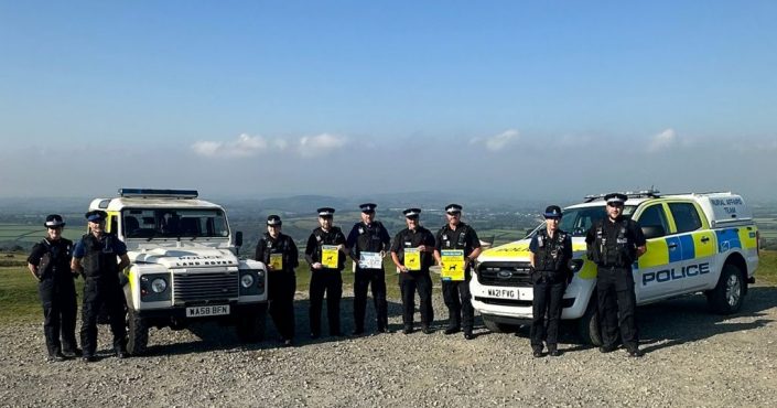 Devon & Cornwall Police officers and PCSOs standing next to a police Land Rover and Rural Affairs vehicle with a view of Dartmoor in the background. The officers are holding signs reminding people to keep their dogs on leads and under control